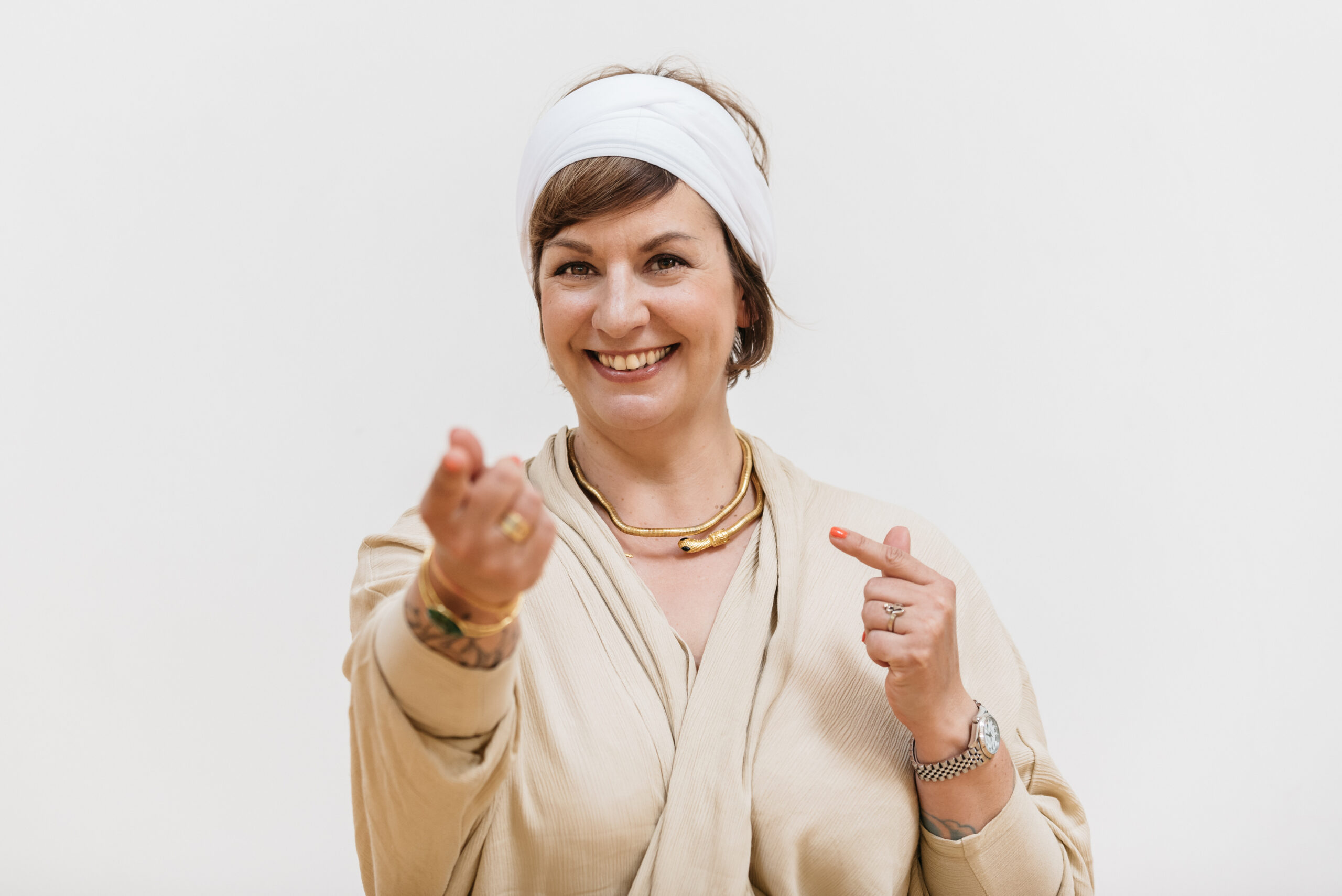 Porträt einer Yogalehrerin vor hellem Hintergrund, lächelnd und mit einladender Handgeste – Ausdruck von Offenheit, Präsenz und achtsamer Begleitung.